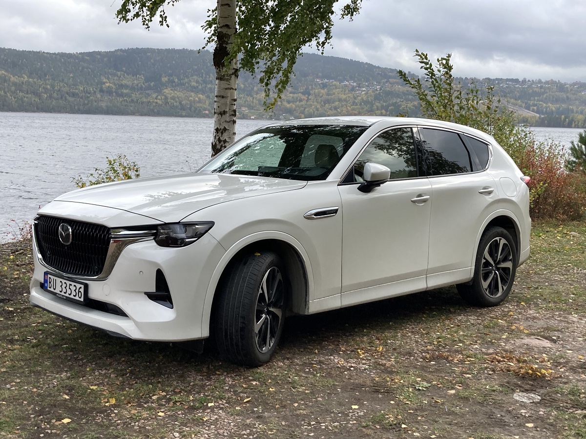 Hvit Mazda CX-60 parkert under en bjørk, foran en innsjø, med høstløv på bakken.