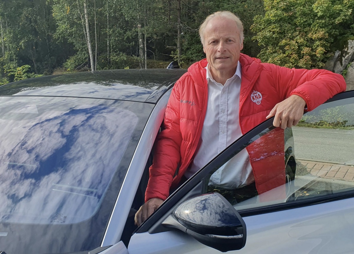 Mann med rød jakke og grått hår, står i døråpnongen på grå bil