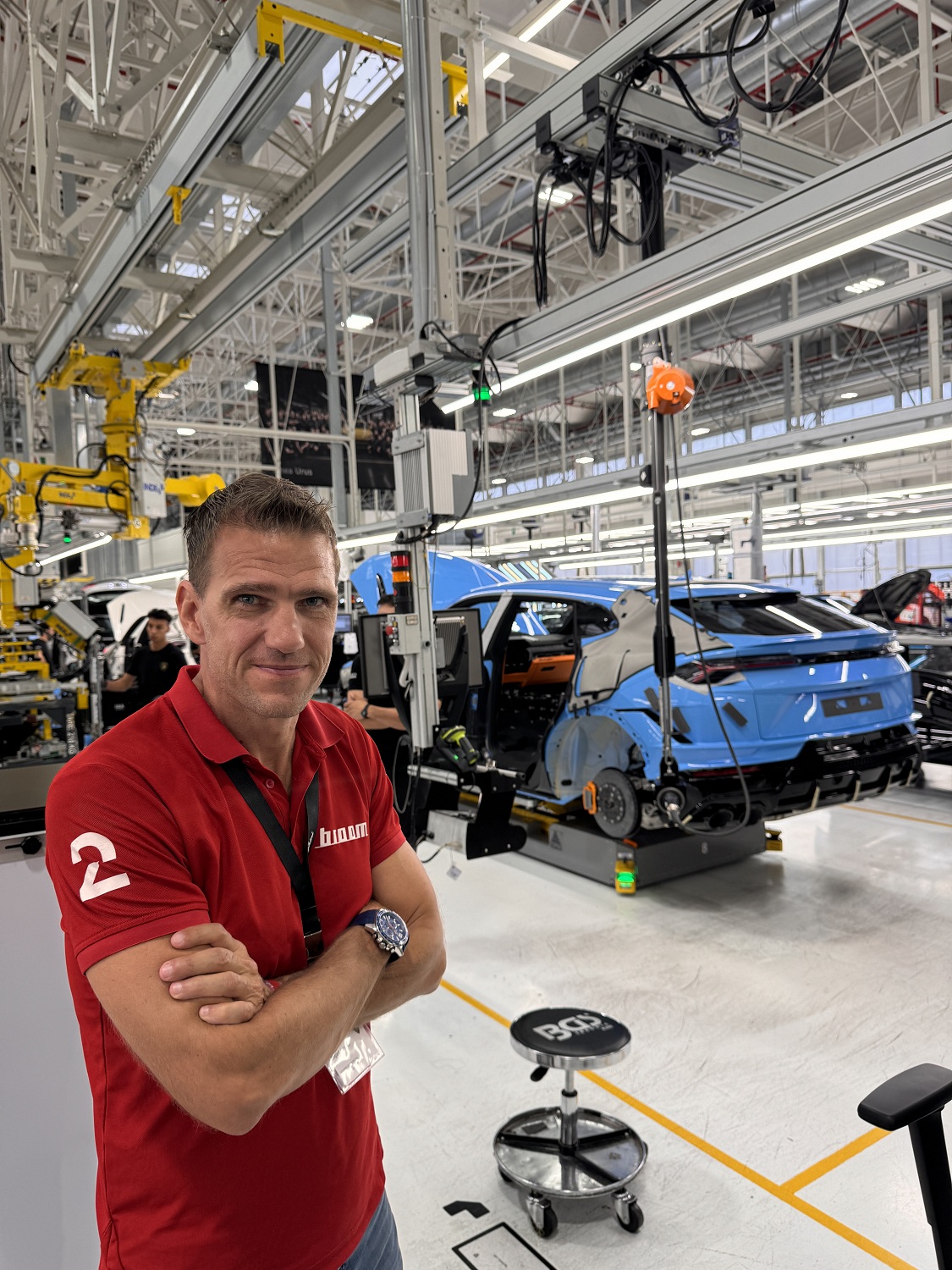 En mann med rød t-skjorte, foran en blå bil som holder på å monteres inne på en bilfabrikk