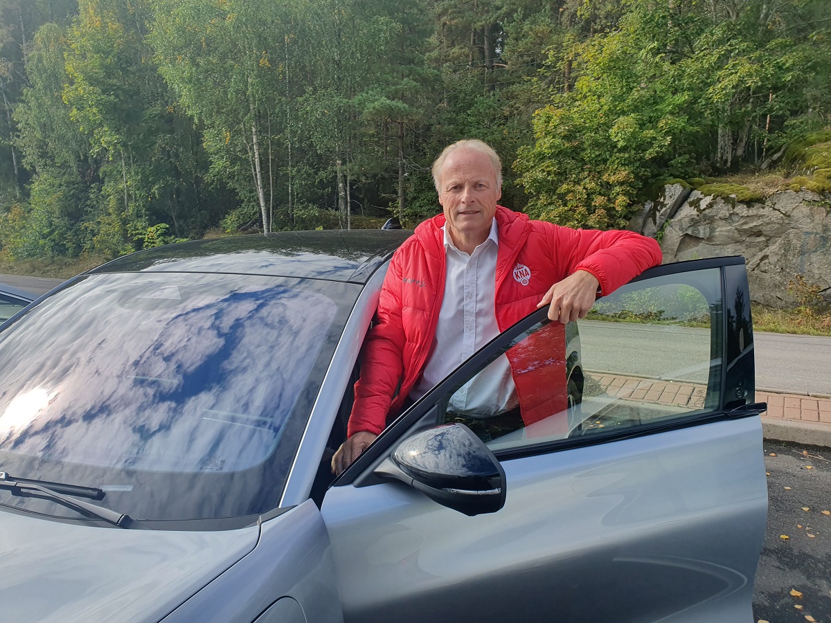 Mann med rød jakke og grått hår, står i døråpnongen på grå bil