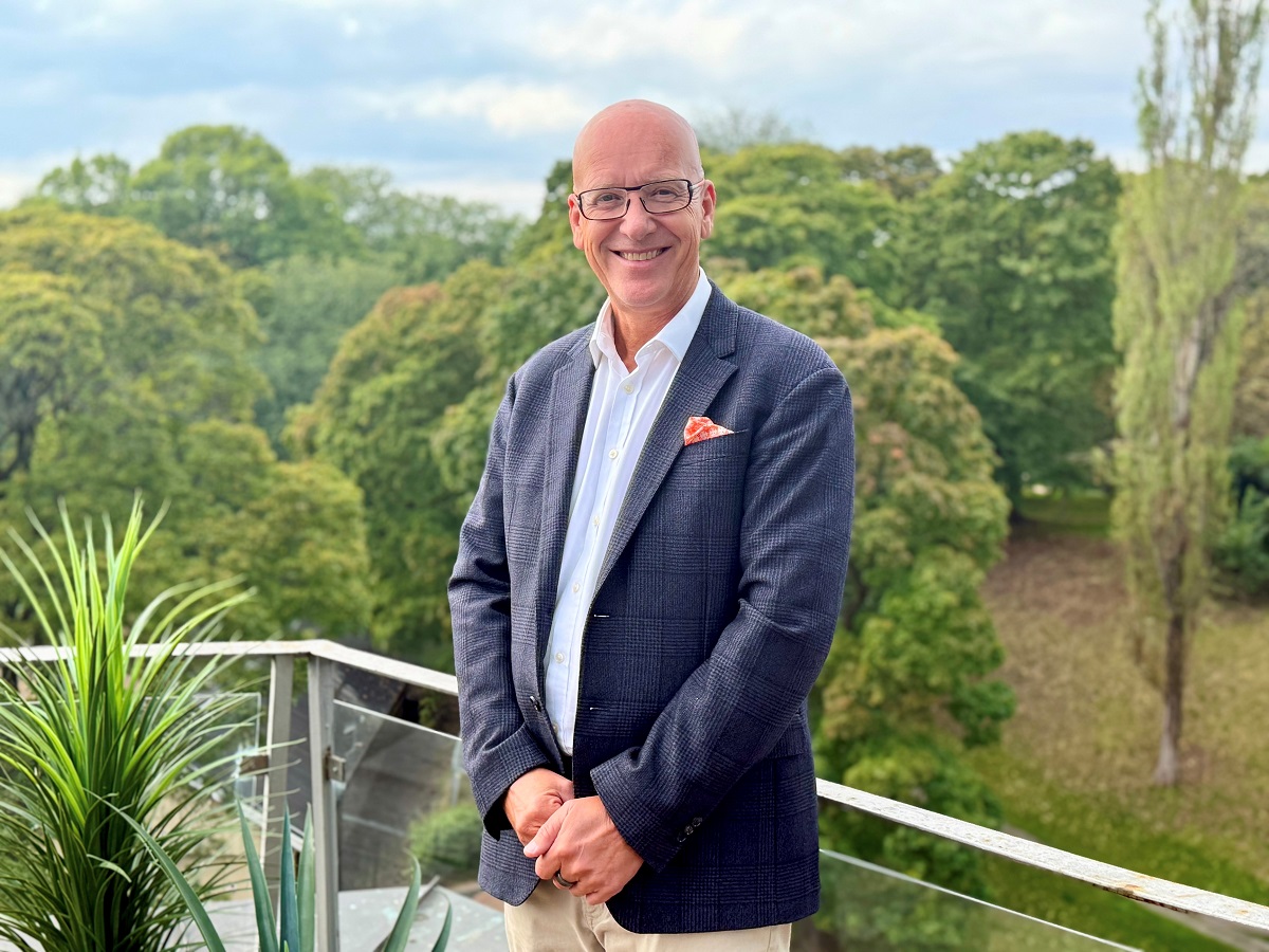 Voksen mann i blå dress på en veranda med glassrekkverk foran grønne lævtrær