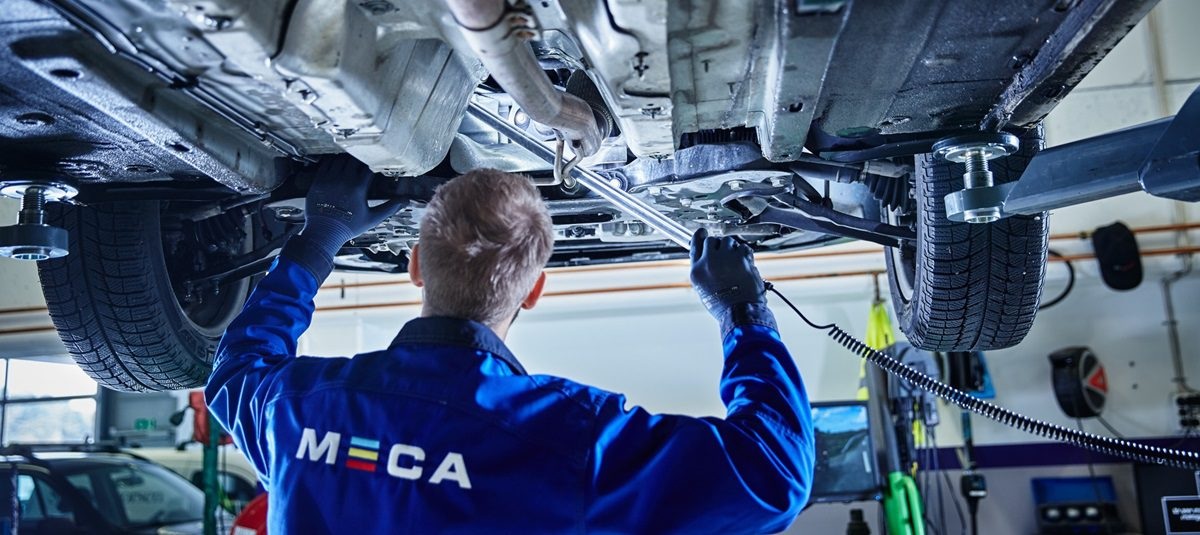 Mekaniker med blå MECA kjeledress og ryggen til, sjekker under bilen